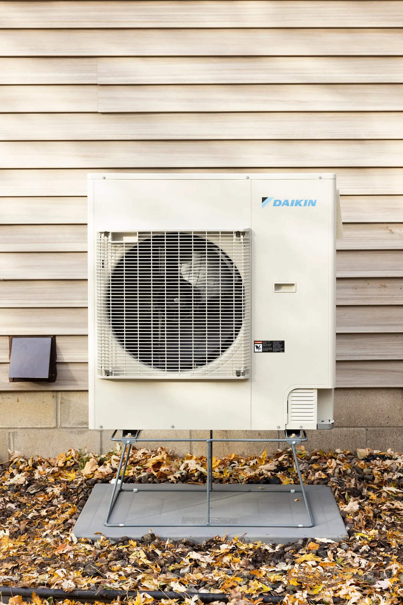 Daikin heat pump outdoor unit mounted on a raised stand against siding, surrounded by fall leaves